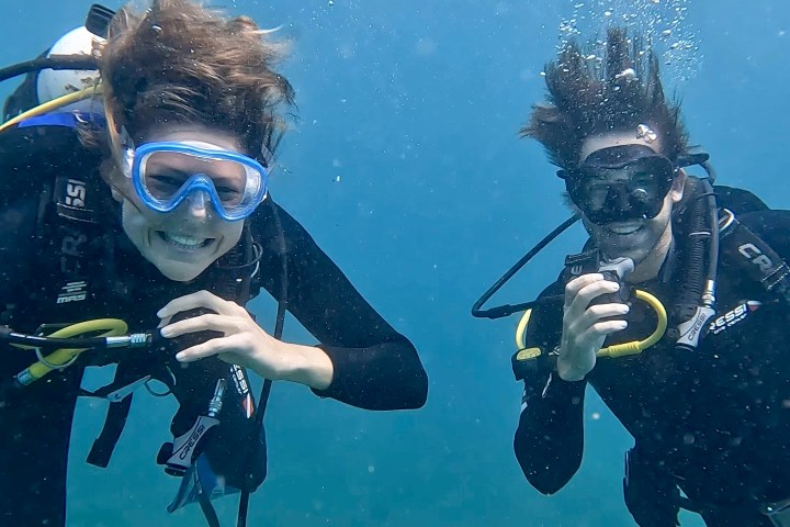 Two scuba divers underwater with clear goggles, wearing wetsuits and gear, surrounded by blue water.