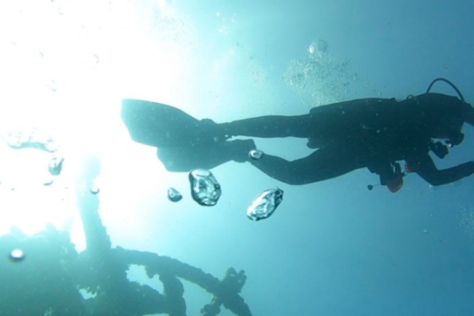 Scuba diver underwater above a submerged structure with sunlight filtering through.