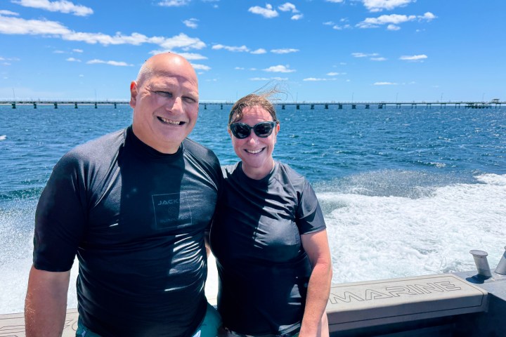Two people in wetsuits on a boat with a bridge in the background.