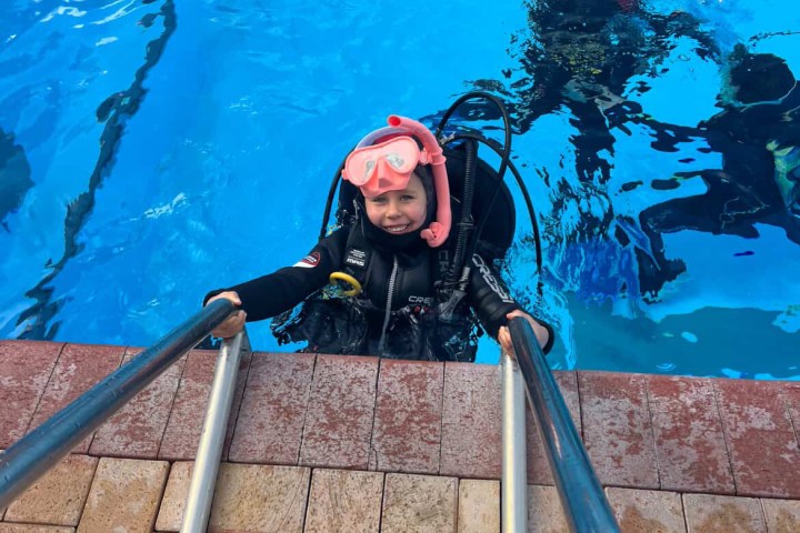 Child in scuba gear climbing out of a pool using a ladder.