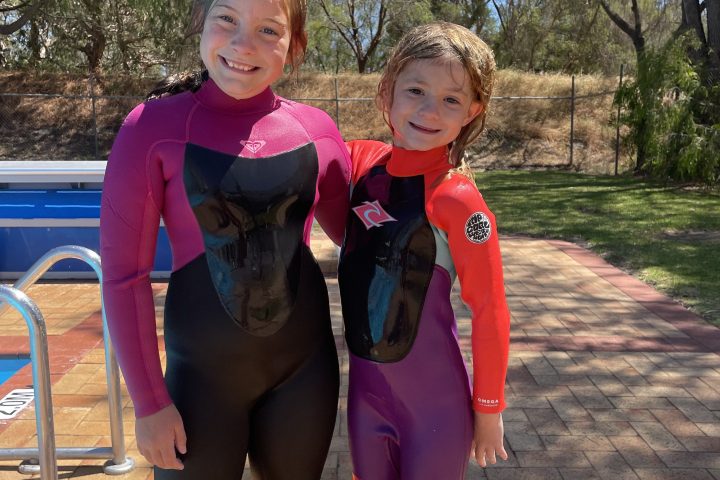Two children in wetsuits standing by a poolside on a sunny day.
