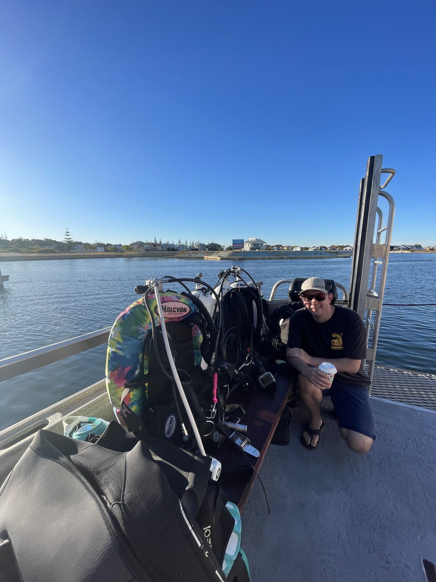 Person crouching on a dock beside scuba gear under a clear blue sky.