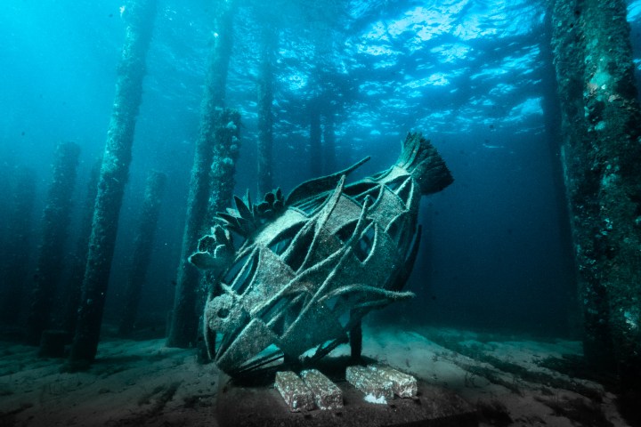 Underwater fish sculpture amidst vertical pillars on the ocean floor.